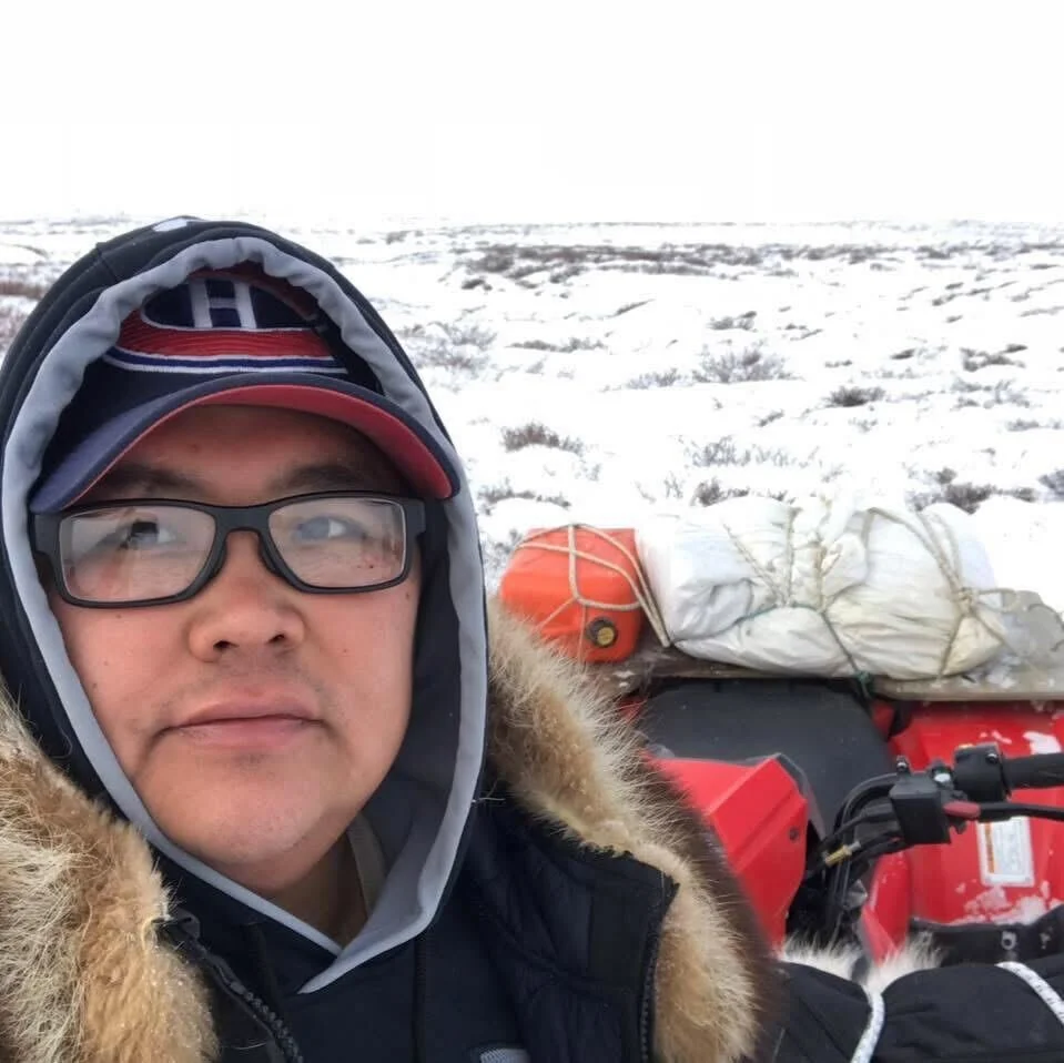 A man wearing glasses, a winter coat with a fur-lined hood, and a baseball cap taking a selfie in a snowy landscape with a red ATV and gear in the background.