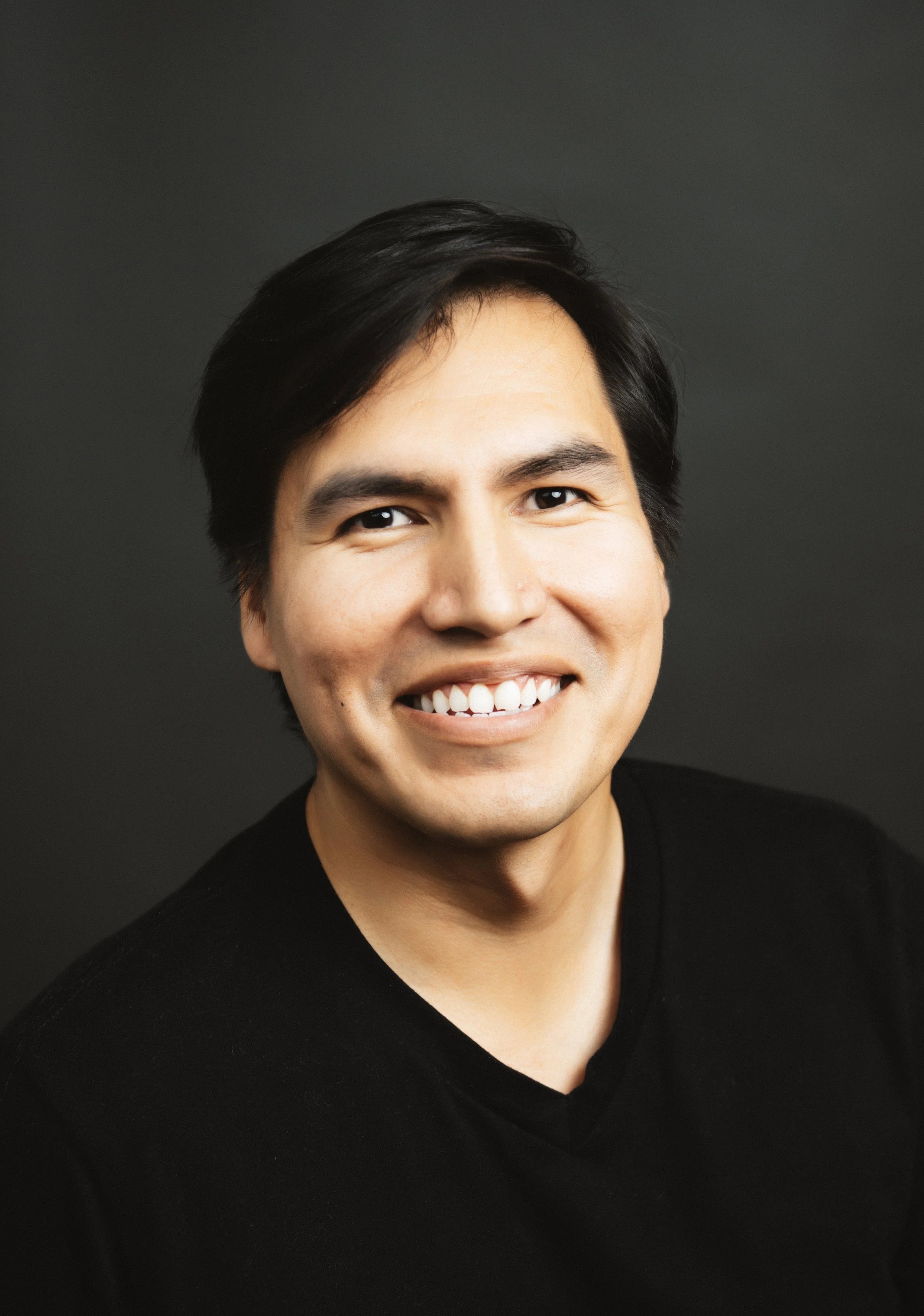Headshot of a man with dark hair, smiling, wearing a black shirt against a dark background.