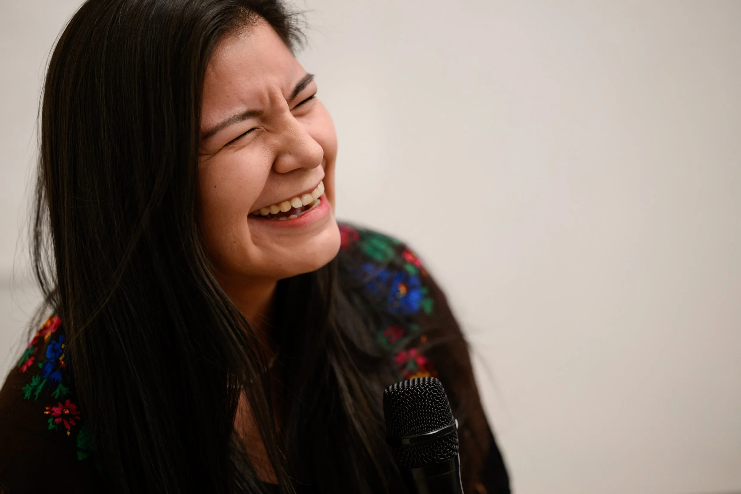 A woman with long dark hair laughing with her eyes closed, holding a microphone, wearing a black top with colorful floral embroidery, against a plain background.