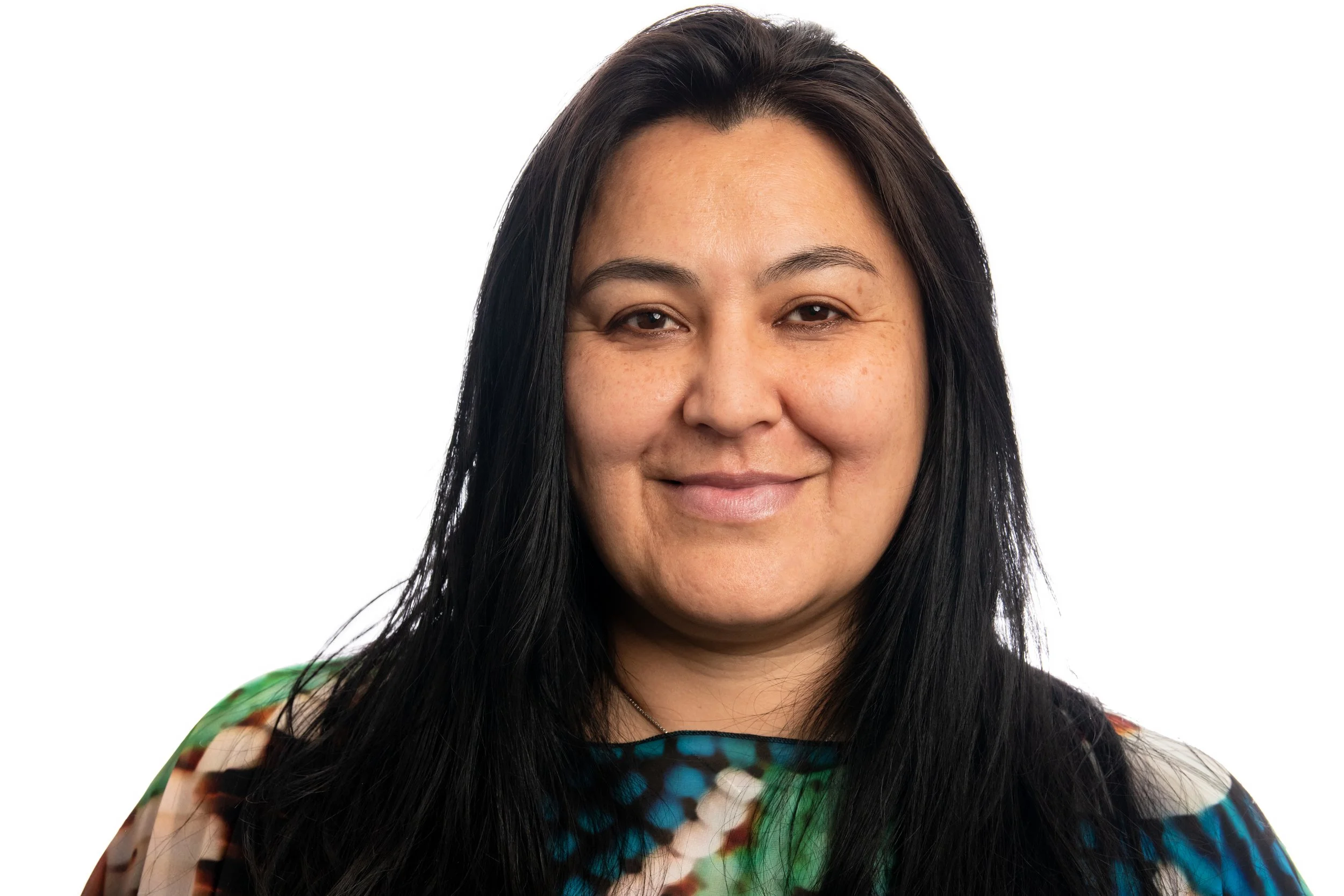 Close-up image of a woman with long black hair, wearing a colorful patterned top, smiling softly against a white background.