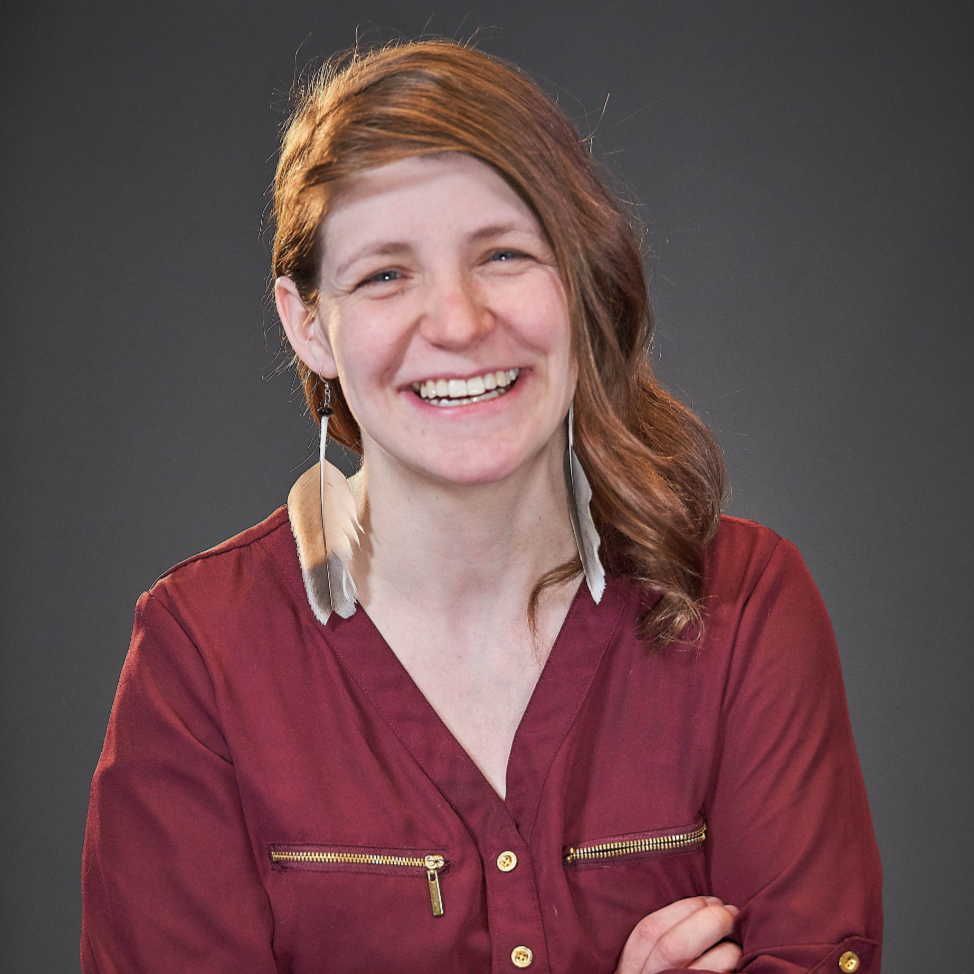 A woman with red hair smiling, wearing a maroon top with gold zippers, against a dark gray background.
