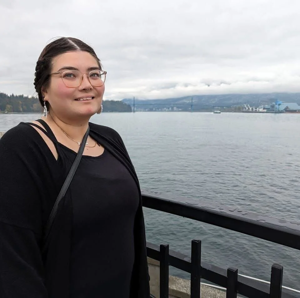 A woman smiling by a waterfront with a bridge and ships in the background on a cloudy day.