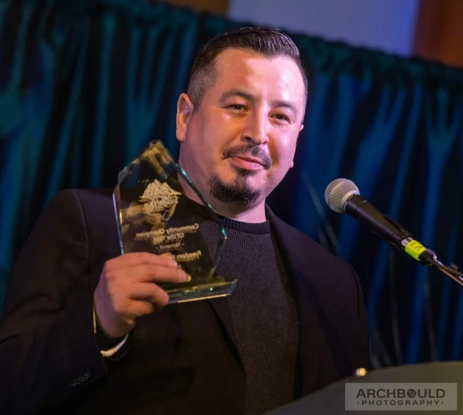 A man holding a glass award or trophy at a ceremony, standing in front of a microphone with a dark curtain background.