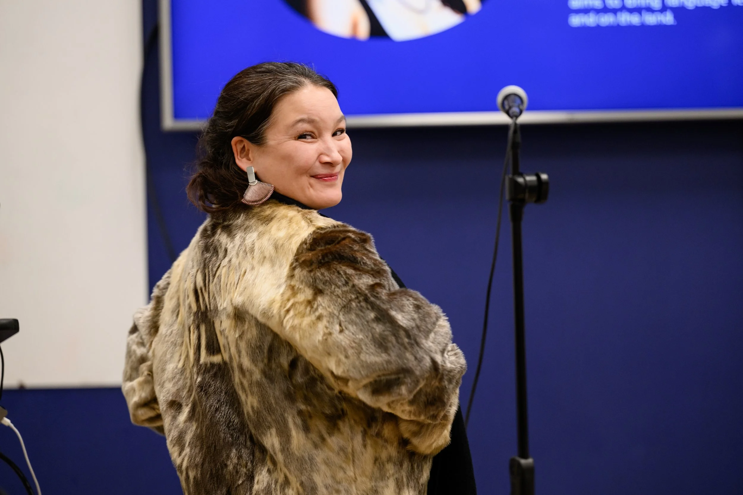 A woman with dark hair smiles and looks over her shoulder, wearing a fur-like coat and large pink earrings, standing in front of a blue background with a microphone nearby.