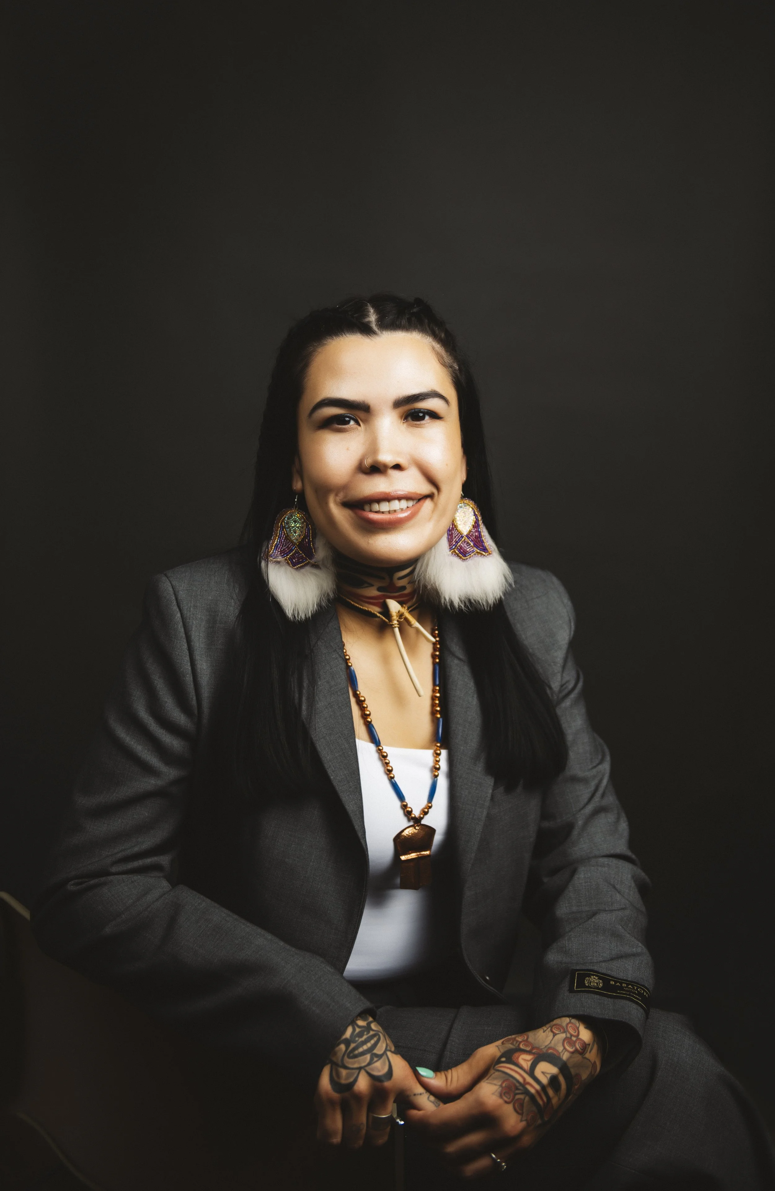 Portrait of a woman with long black hair, tattoos on her hands, wearing a gray blazer, colorful earrings, and layered necklaces, sitting against a dark background.