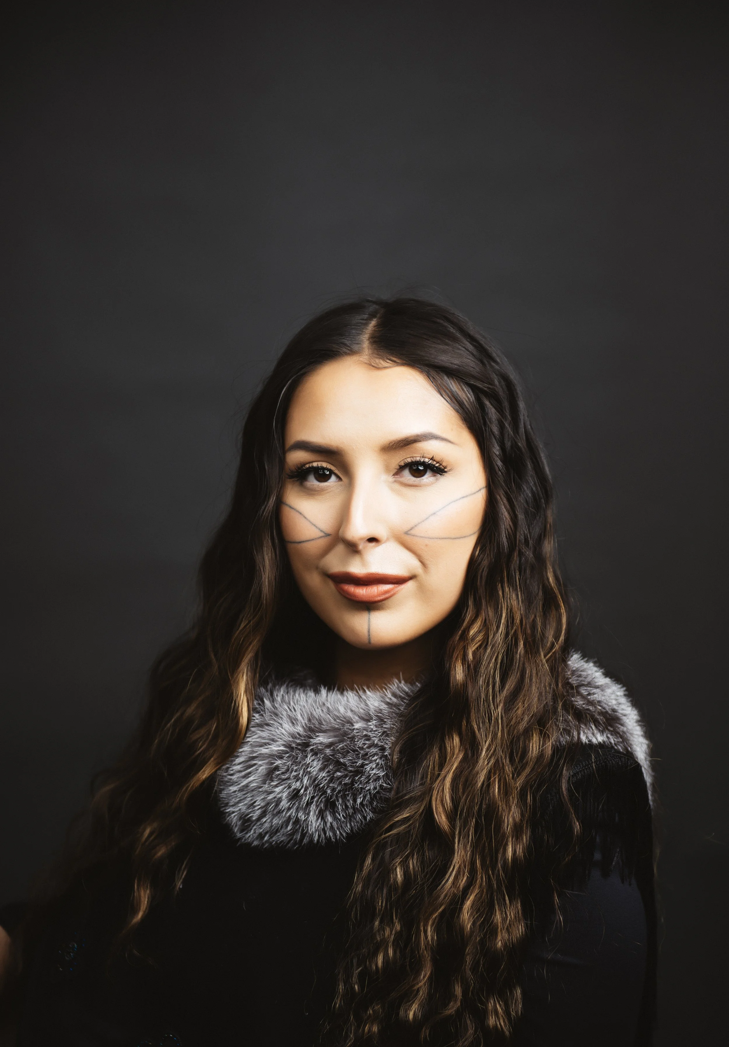 A woman with long, wavy brown hair and makeup, wearing a black outfit with a gray fur collar, has lines drawn on her face against a dark background.