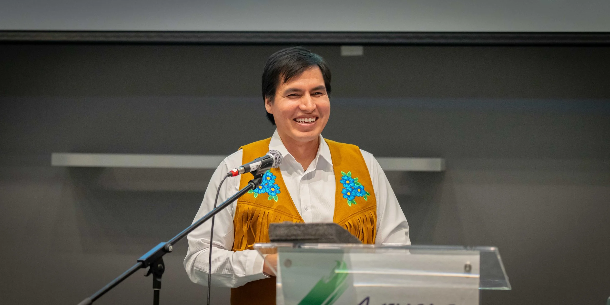 A man with dark hair, wearing a white shirt and a brown vest with blue embroidered flowers, stands behind a clear lectern, smiling and speaking into a microphone.