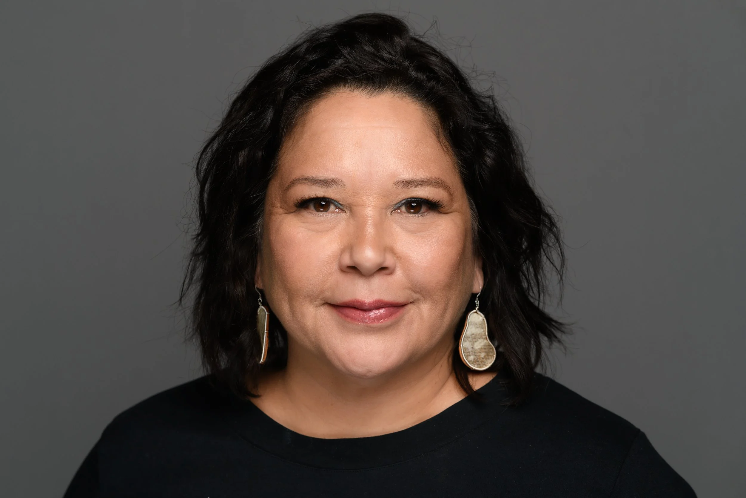 A woman with dark, wavy hair and hoop earrings smiling at the camera, wearing a black top against a gray background.