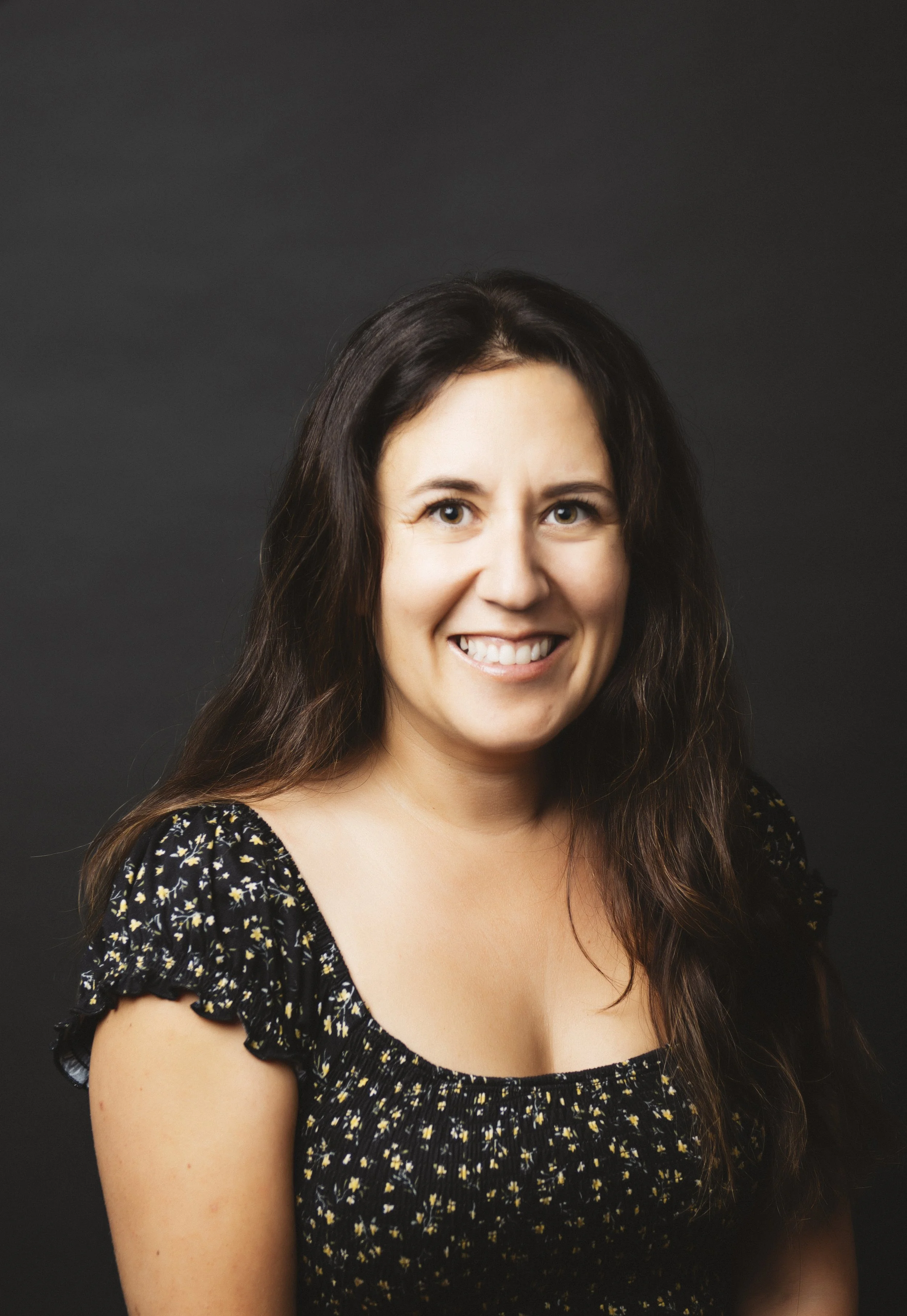 A woman with long dark hair smiling at the camera, wearing a black floral dress, against a plain dark background.