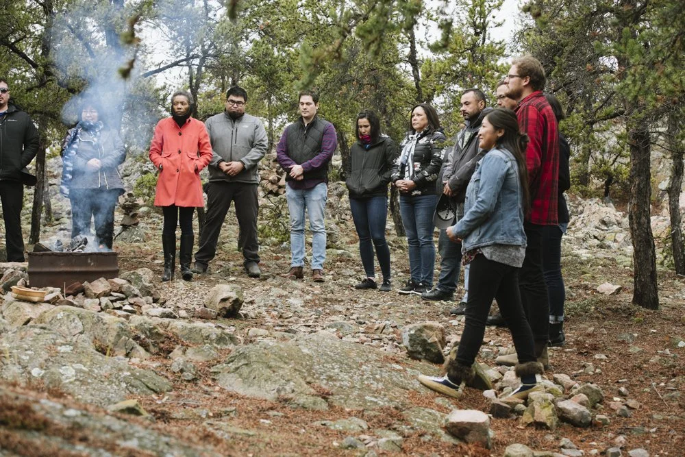 Group of people standing outdoors in a forested area, gathered around a smoking firepit.