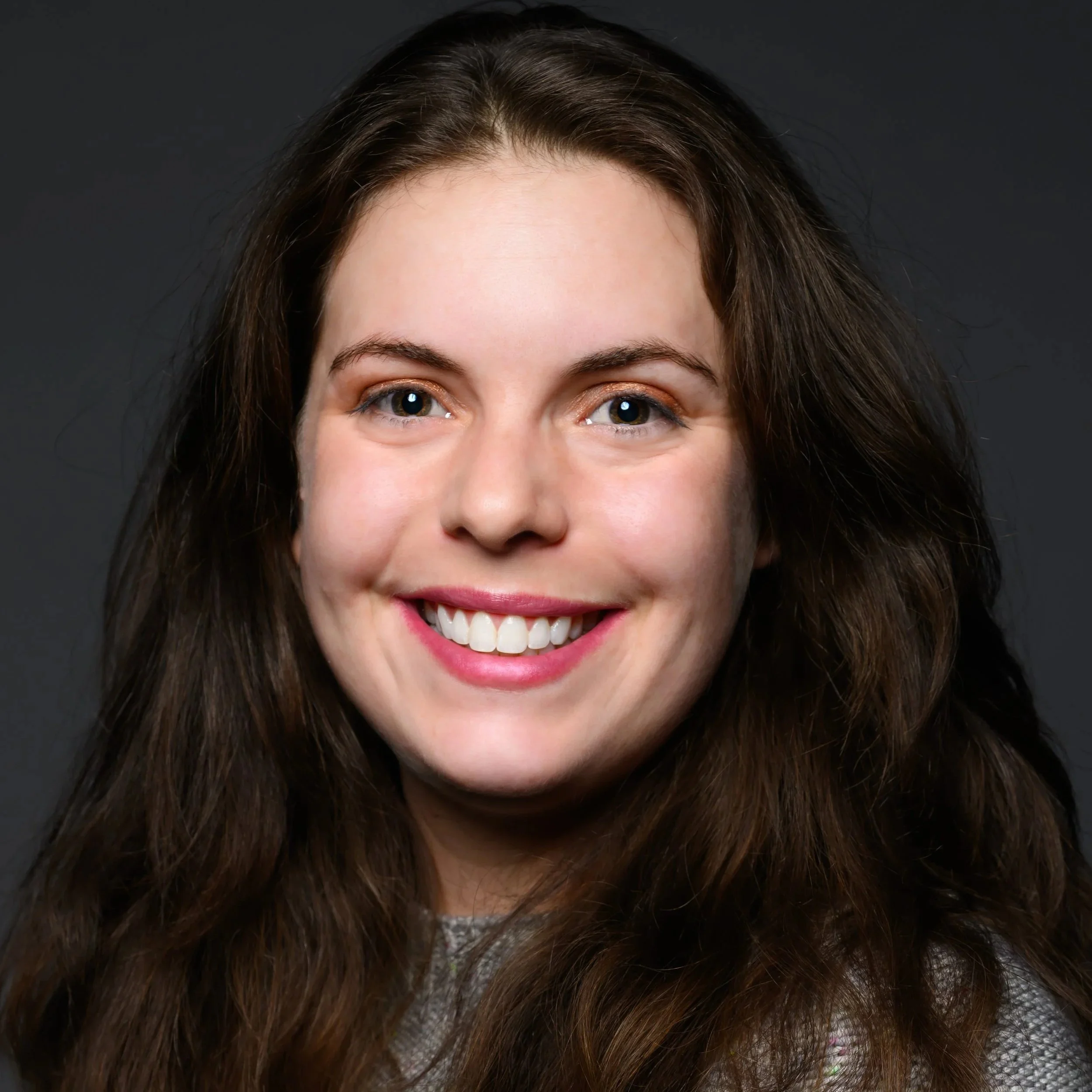 A young woman with long, wavy brown hair smiling against a dark background.
