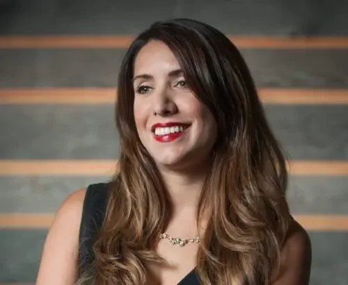 A woman with long, wavy brown hair and red lipstick smiling, wearing a black sleeveless top and a necklace against a wooden background.