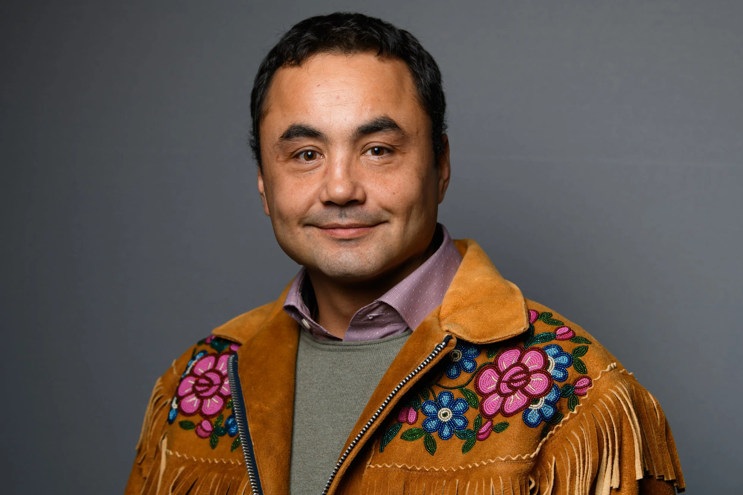 A man with dark hair, wearing a brown embroidered fringed jacket and a light purple collared shirt, smiling against a plain gray background.