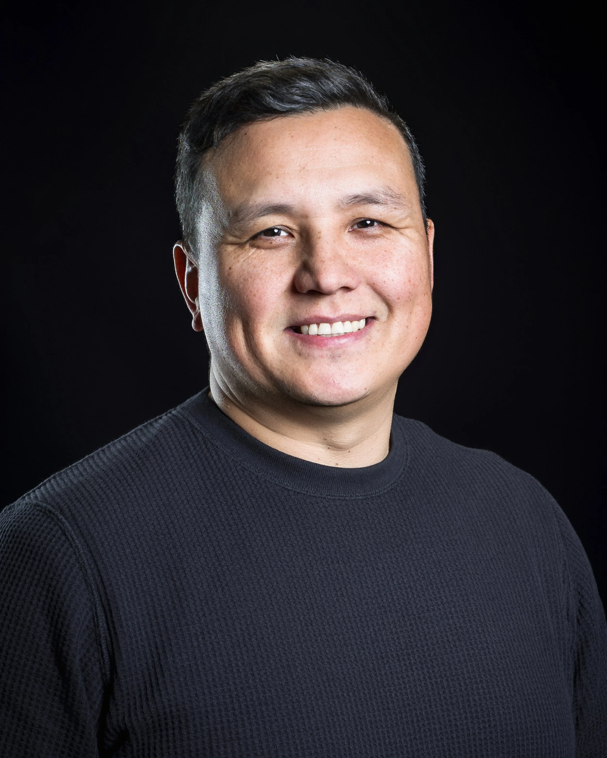 Portrait of a smiling man with short dark hair, wearing a black textured shirt, against a dark background.