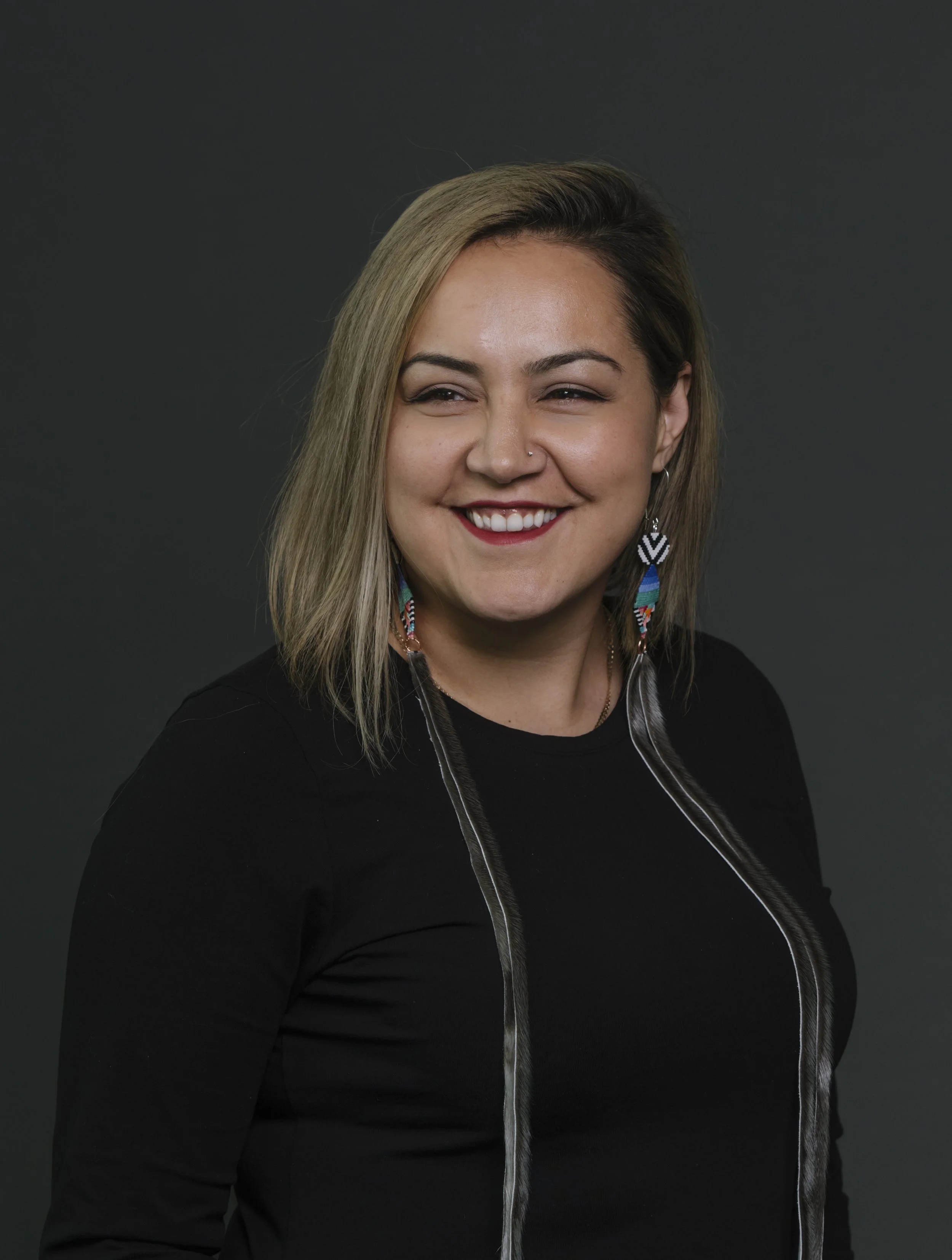 A woman with shoulder-length blonde hair wearing a black top and colorful beaded earrings, smiling against a dark background.