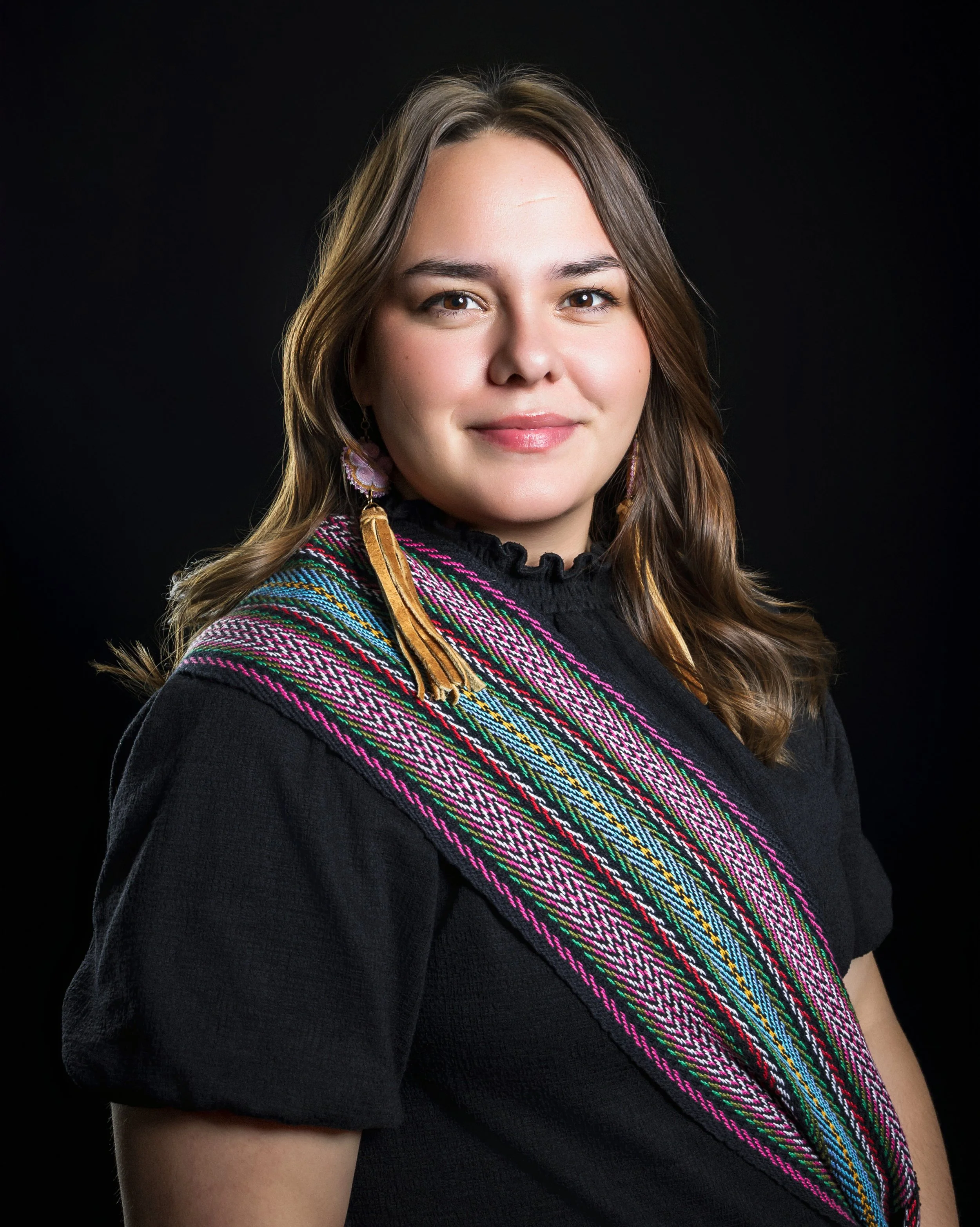 A woman with light skin and long, wavy brown hair wearing a black top and a colorful woven shawl, standing against a black background.
