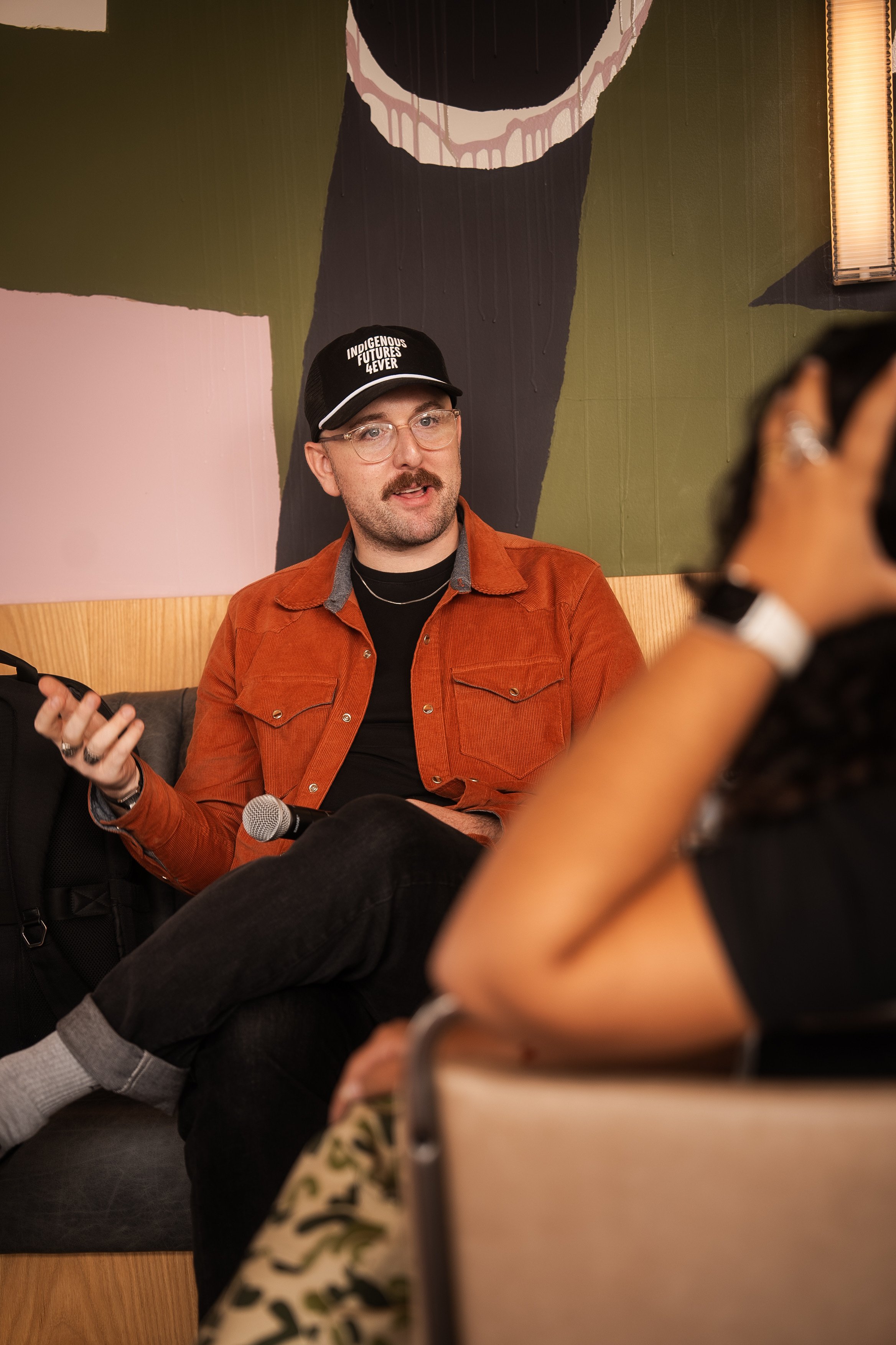A man wearing a black cap, glasses, and an orange jacket sitting on a couch, speaking with a woman in a black shirt in an indoor setting with a colorful mural on the wall behind him.