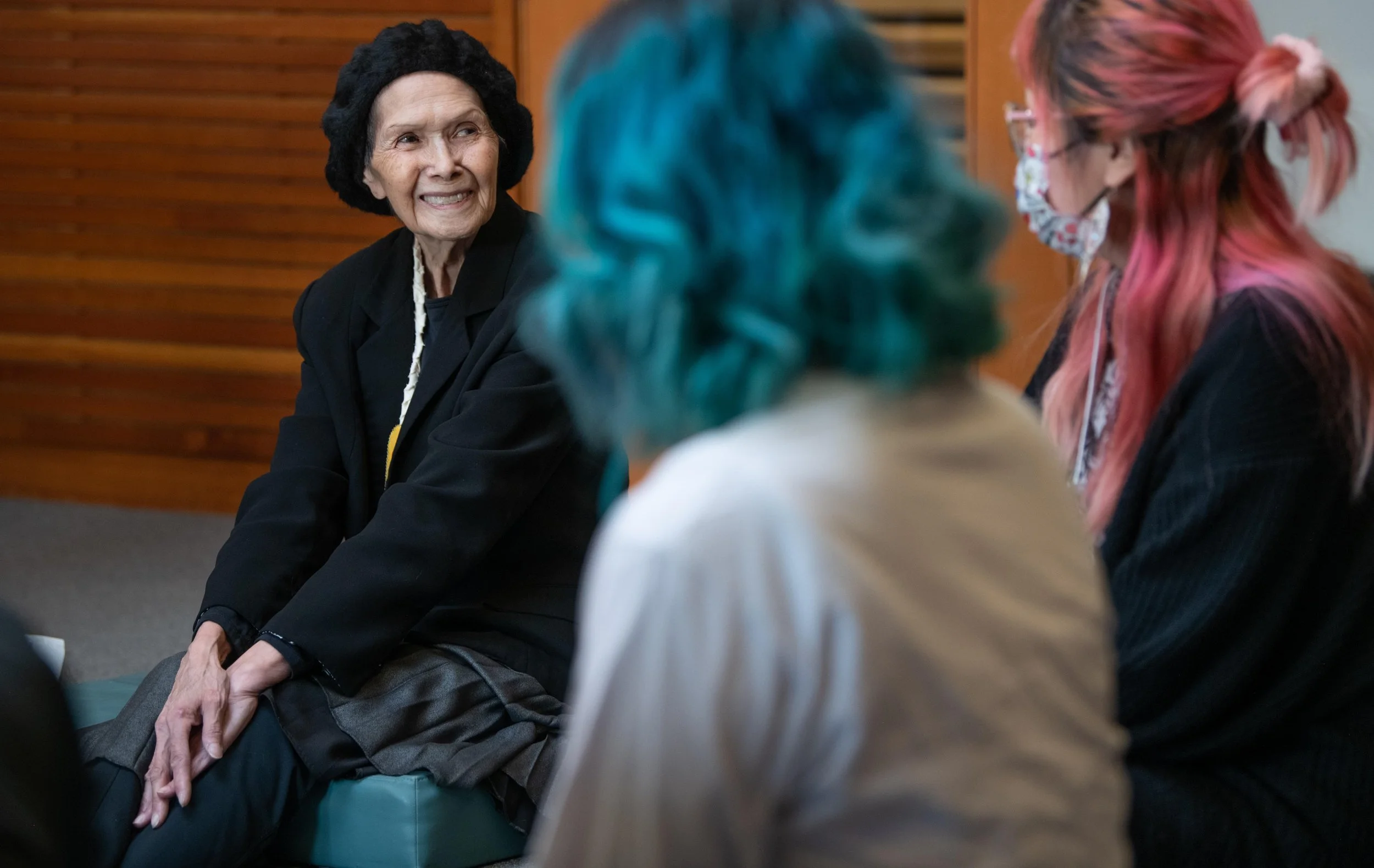 An elderly woman with a black hat and black blazer smiling and engaging in a conversation with two women with colorful hair, one with blue and the other with pink, in a room with wooden walls.