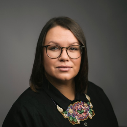Portrait of a woman with glasses wearing a black top and a colorful beaded necklace against a neutral background.