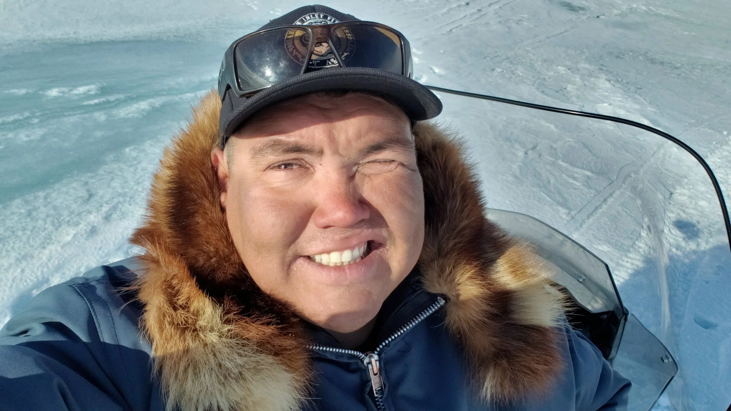 A man taking a selfie outdoors on a snow-covered landscape, wearing a winter jacket with a fur-lined hood, a cap, and sunglasses on his head.