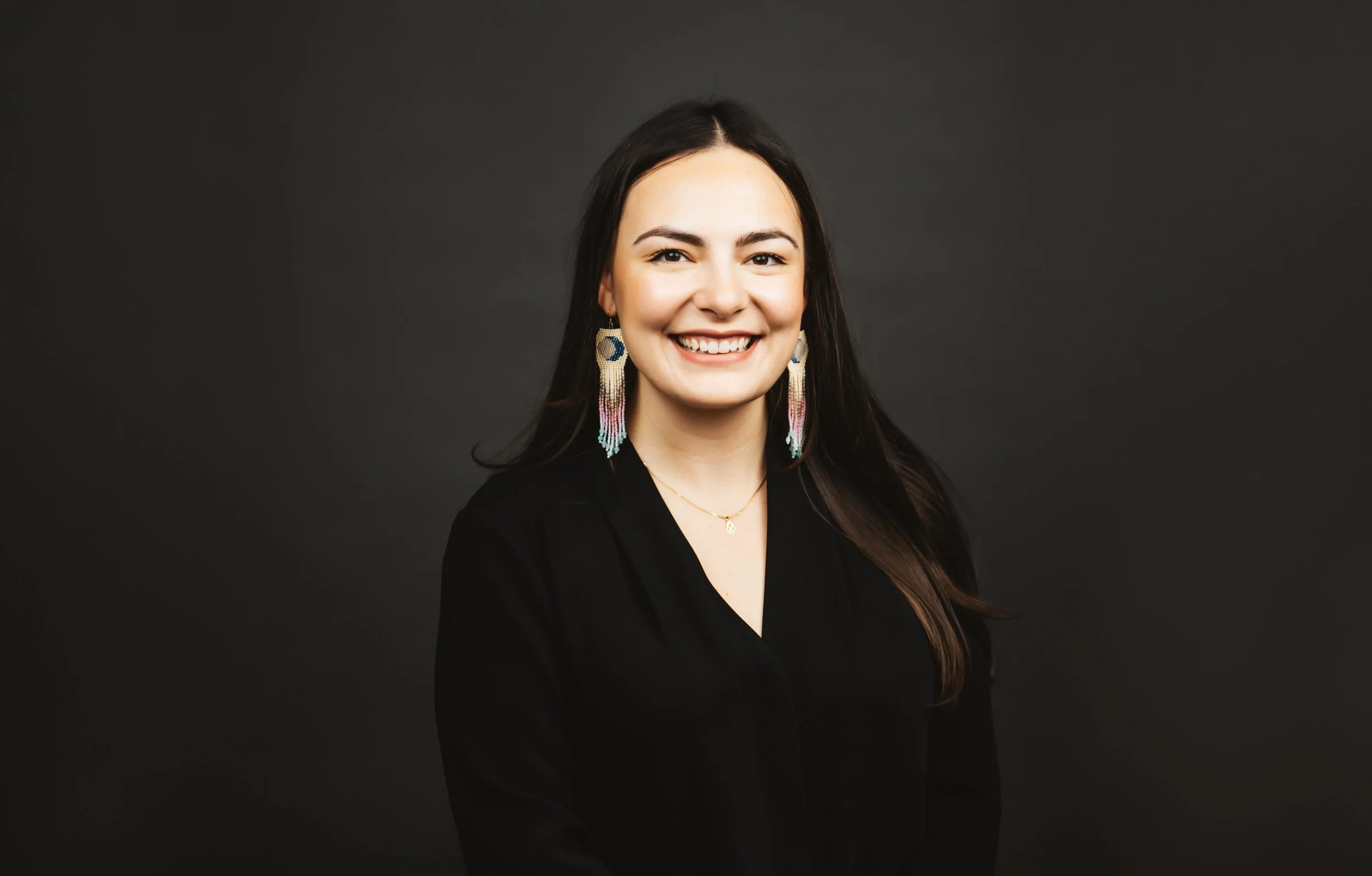 A smiling woman with dark hair, wearing colorful earrings, a black top, and a delicate necklace, against a dark gray background.