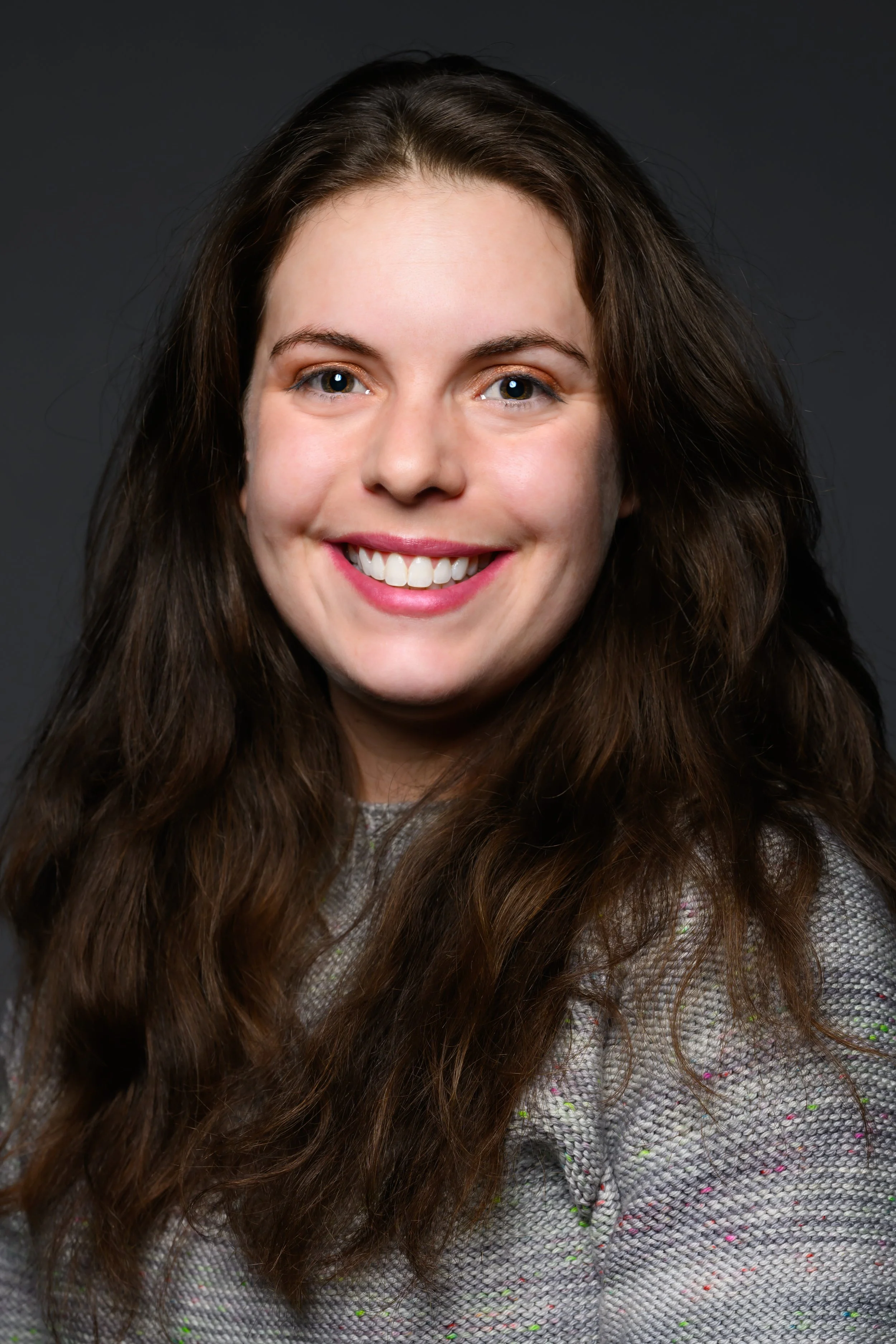 A young woman with long, wavy brown hair smiling at the camera, wearing a grey textured top with colorful specks on a dark background.