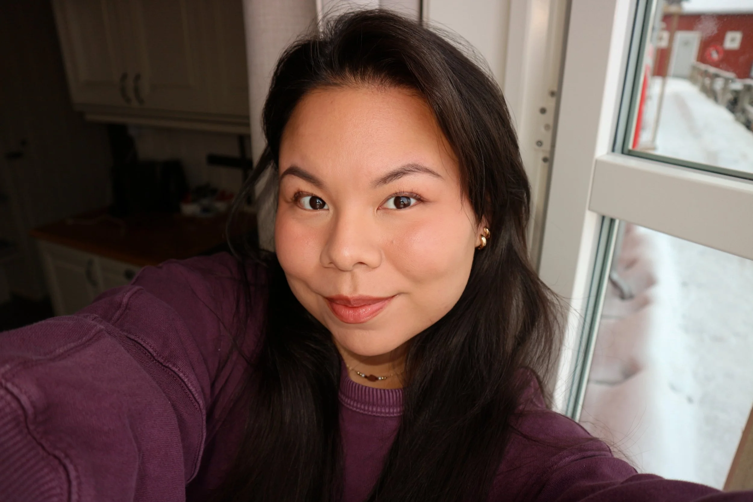 A woman with long dark hair and earrings smiling at the camera, standing near a window with a snowy outdoor scene visible.