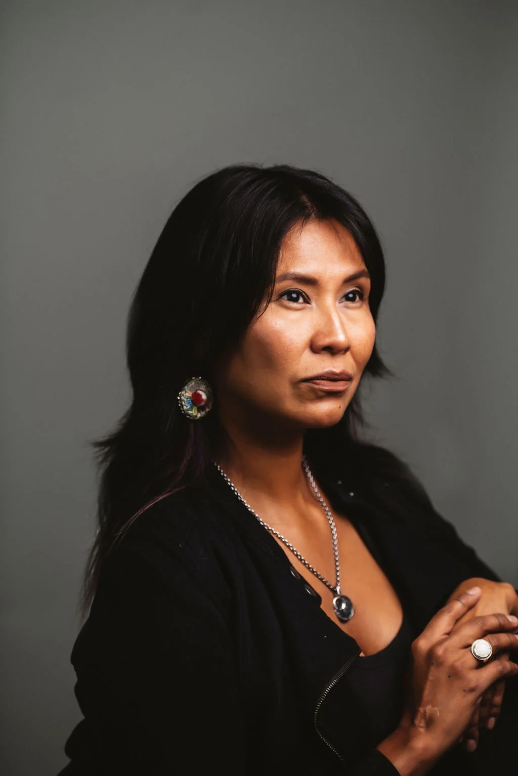 A woman with dark hair, wearing earrings and a necklace, posing against a gray background.