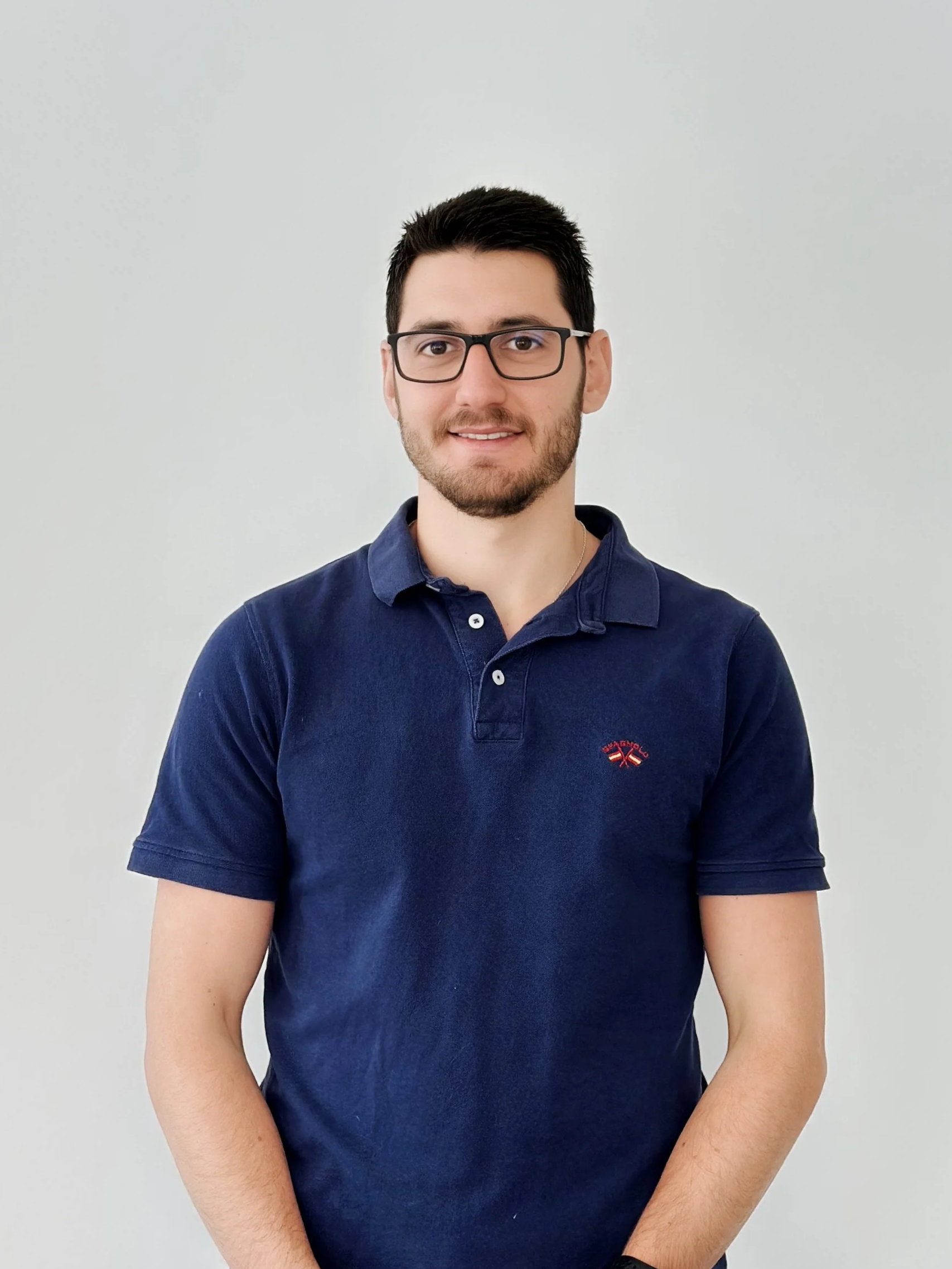 A young man with dark hair, glasses, and a beard, wearing a navy blue polo shirt, standing against a plain white background.