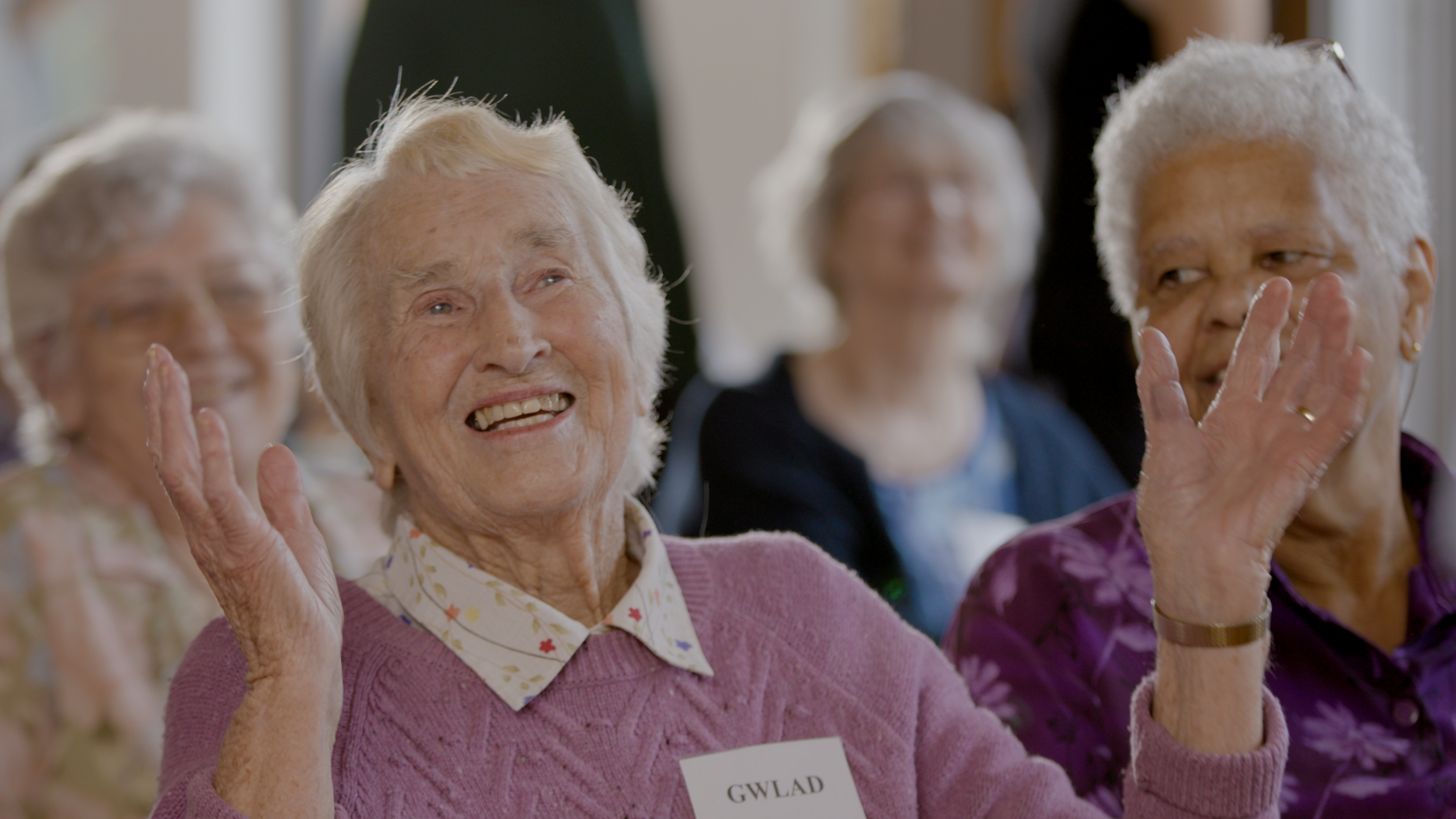 Gwlad, an older woman with fair skin and white hair smiles, while gesturing with her hands in the air. She is surrounded by friends - members of the Rewire Musical Memories Choir.