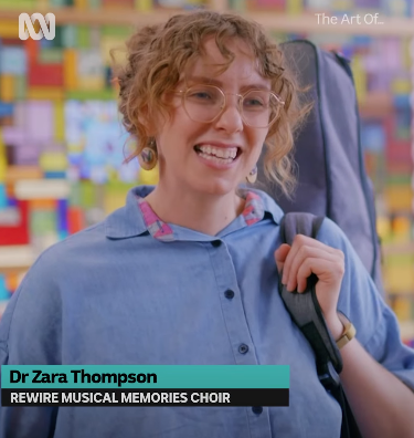 Zara, a white woman with blond hair, is standing with a guitar over her shoulder