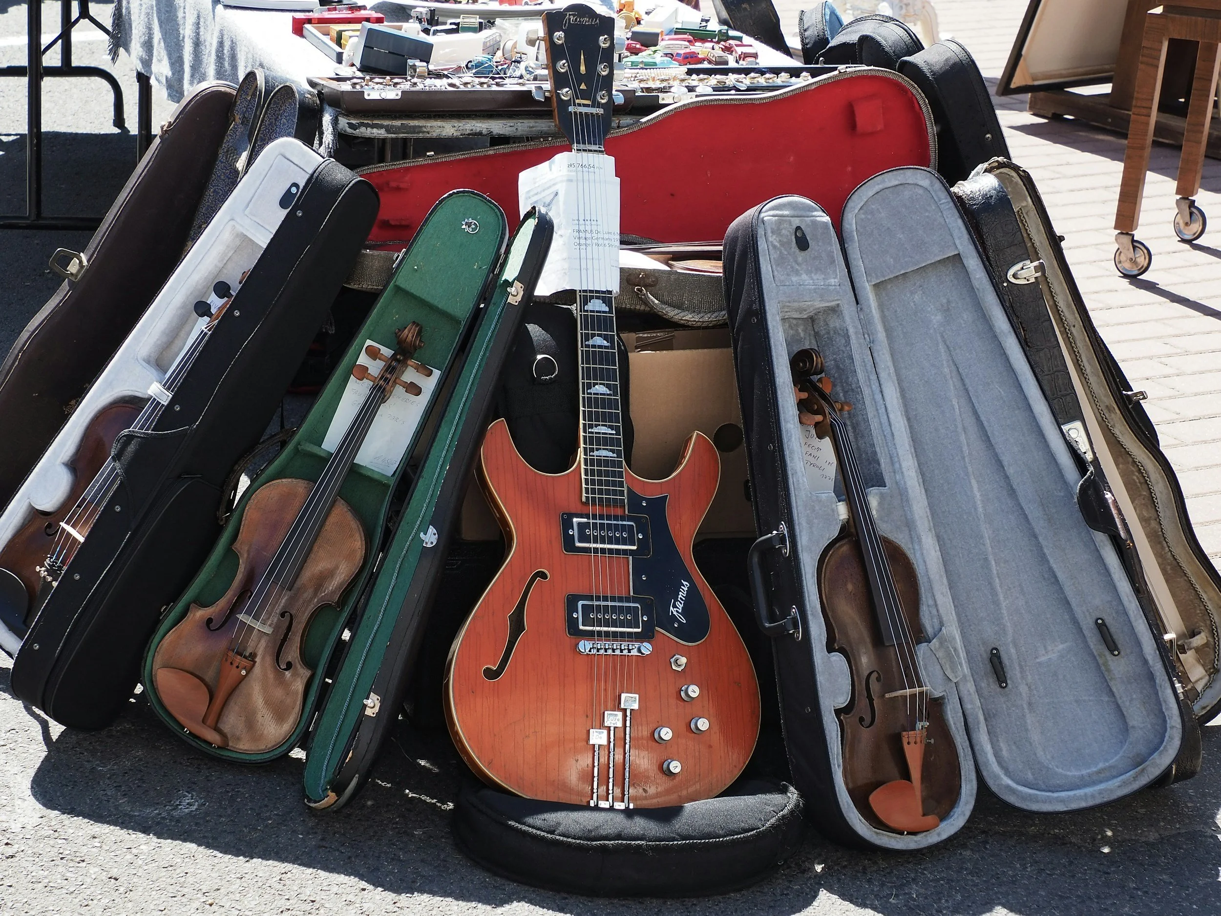 Multiple violins in cases and a semi-hollow electric guitar on a table at an outdoor market.