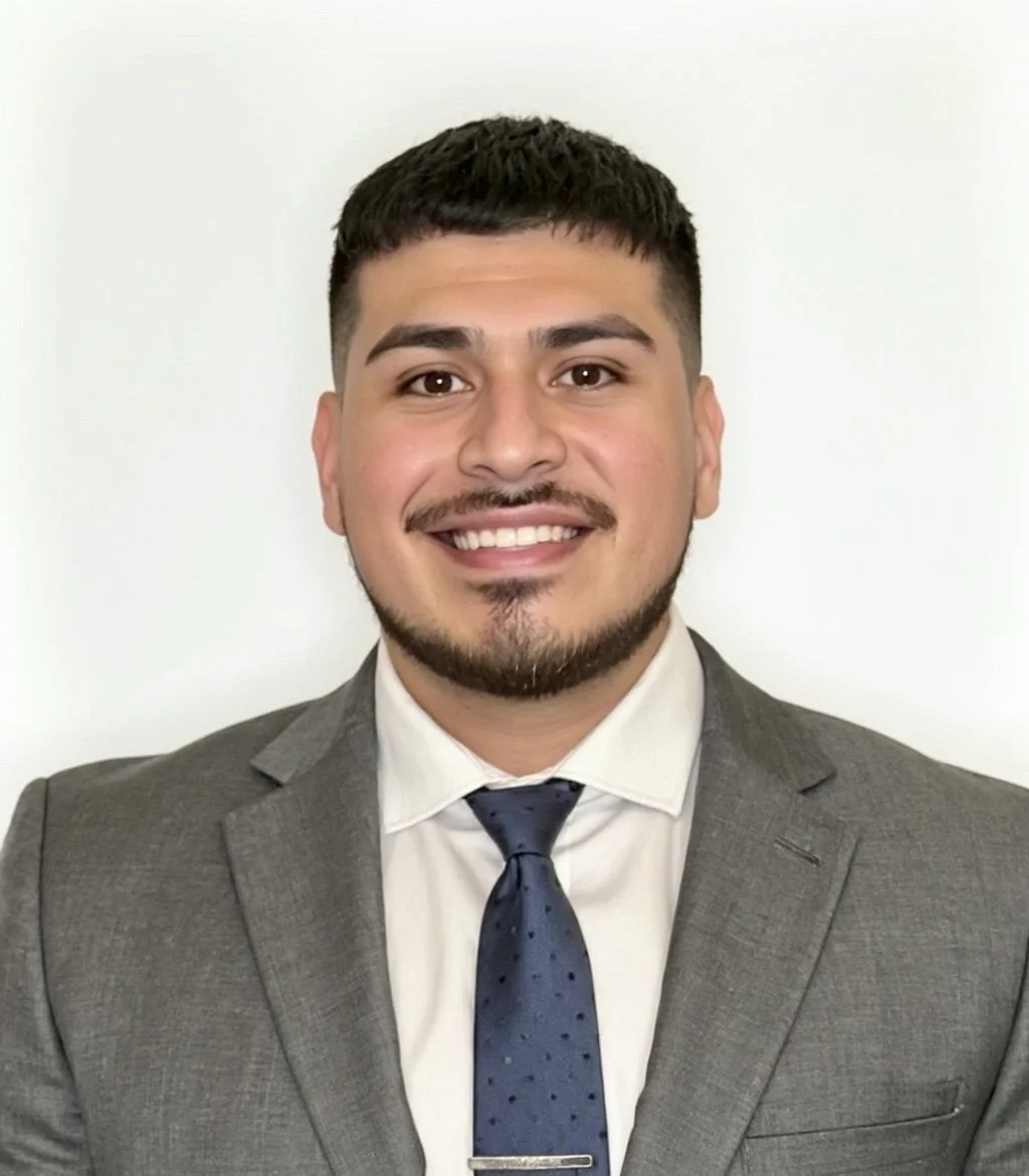 A man with short dark hair, a trimmed beard, wearing a gray suit, white shirt, and a dark blue tie, smiling in front of a plain white background.