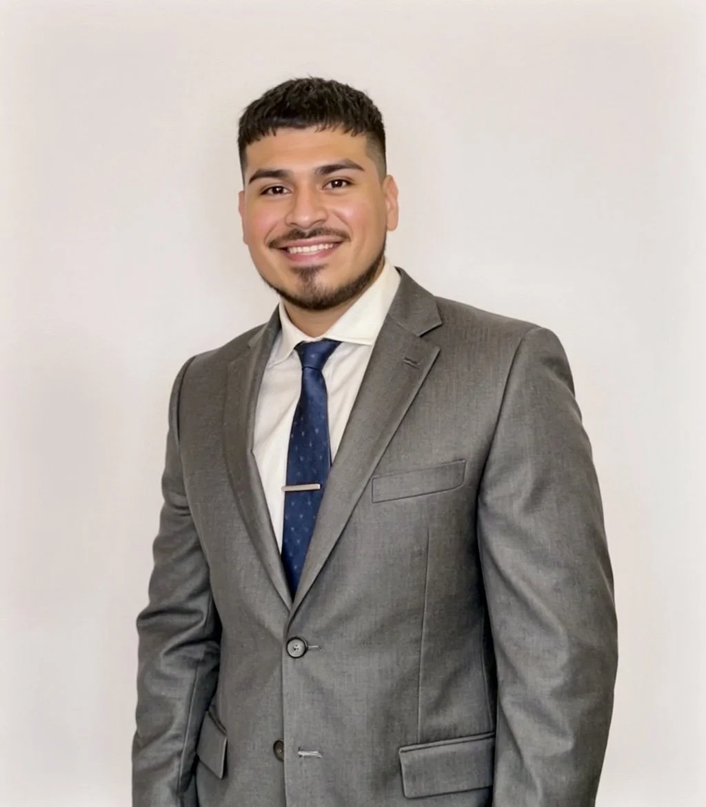 A man in a gray suit, white shirt, and blue tie standing against a plain white wall, smiling at the camera.
