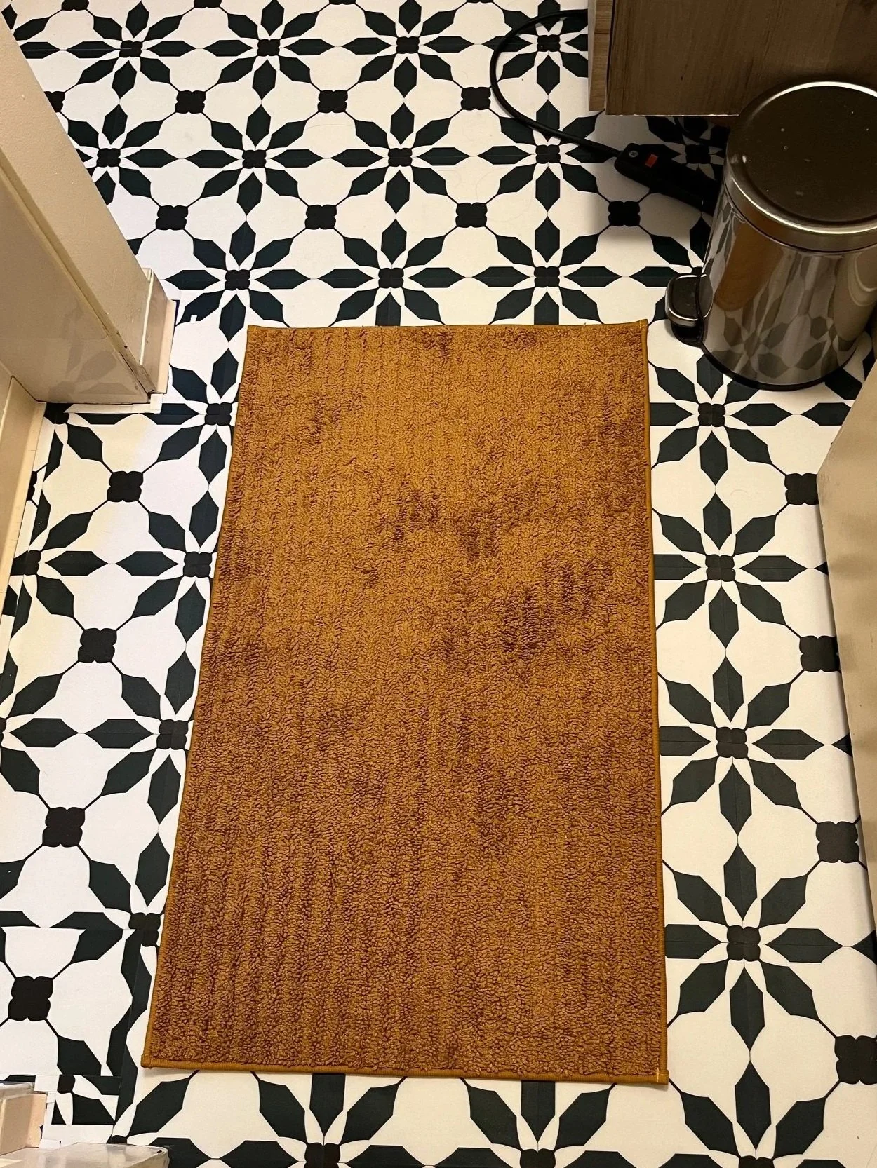 A top-down view of a kitchen floor with black and white geometric tile pattern, a rectangular brown rug, and a black trash can.