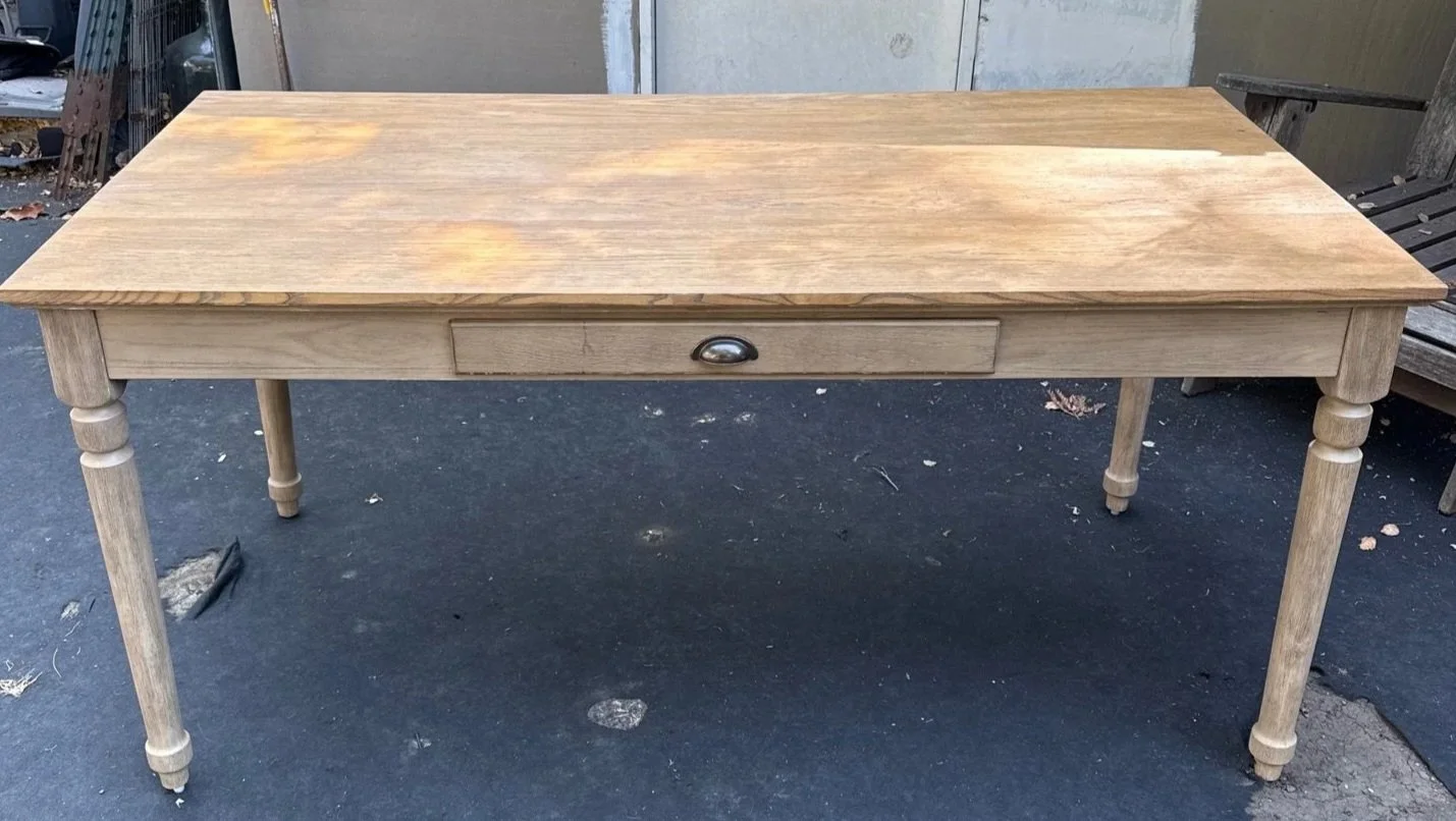 Unfinished wooden table with tapered legs and a single drawer in the front, outdoors on a paved surface.