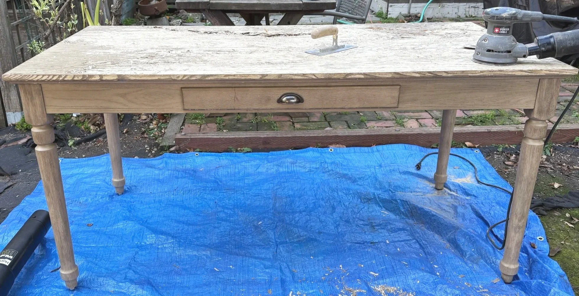 A wooden table with a drawer and turned legs outdoors, resting on a blue tarp with tools and electrical cords nearby.
