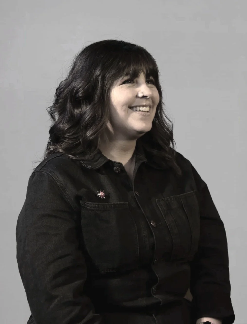 A woman with dark wavy hair smiling, wearing a black denim jacket with a pink and silver pin, sitting against a plain light grey background.