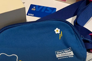 Blue bag with a sticker that reads 'She's Next in fashion' and a VISA card sticking out, positioned near the bag's zip.