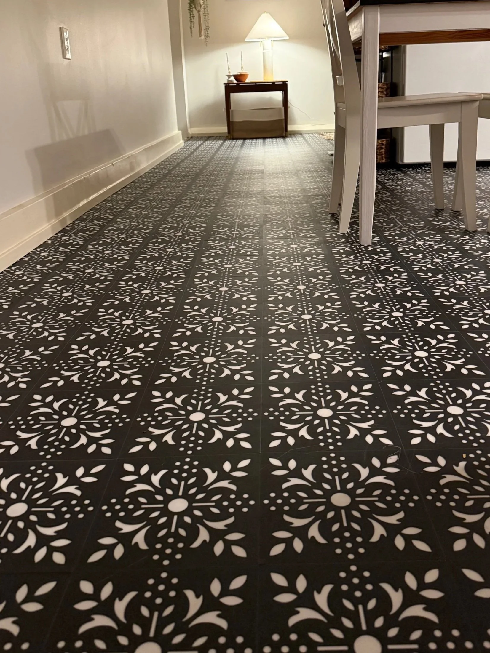 Interior view of a room with patterned black and white floor tiles, a white wall, a small wooden table with a lamp and decorative items, and a dining table with chairs.