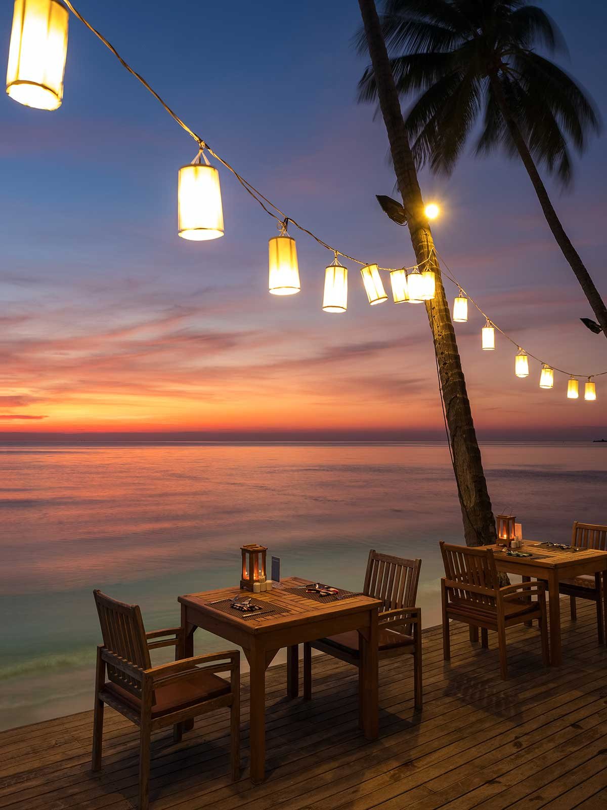 A sunset over the ocean with a wooden deck, two tables with chairs, tropical palm trees, and string lights hanging overhead.