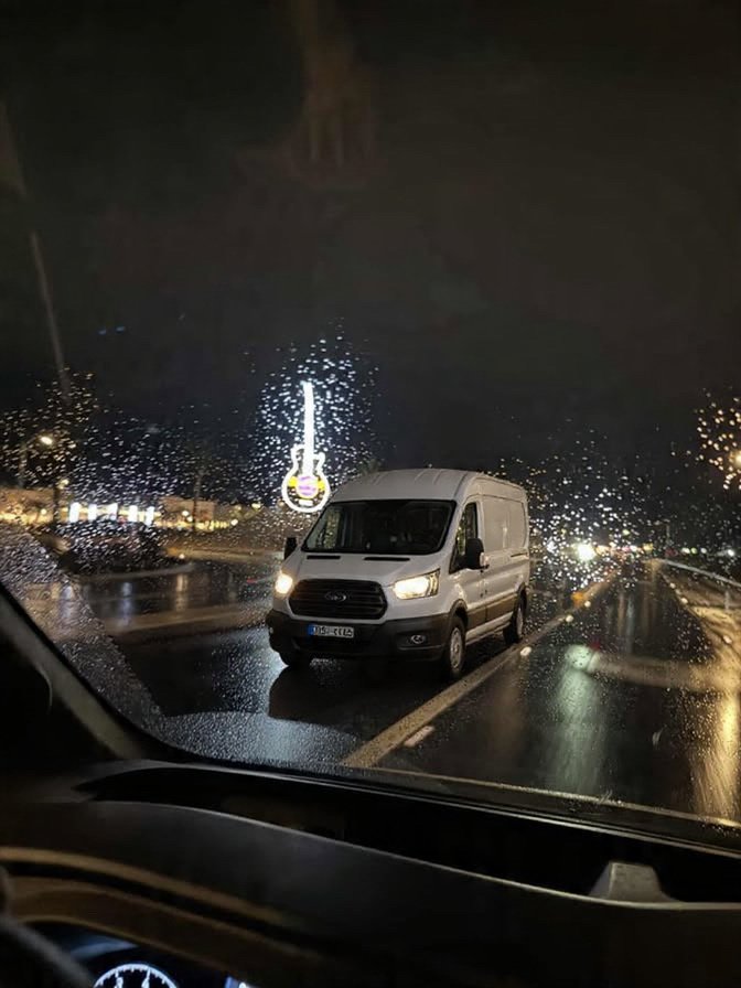 Nighttime rain-streaked car windshield with raindrops on glass, view of a white van on a wet road with blurred city lights and a neon sign in the background.