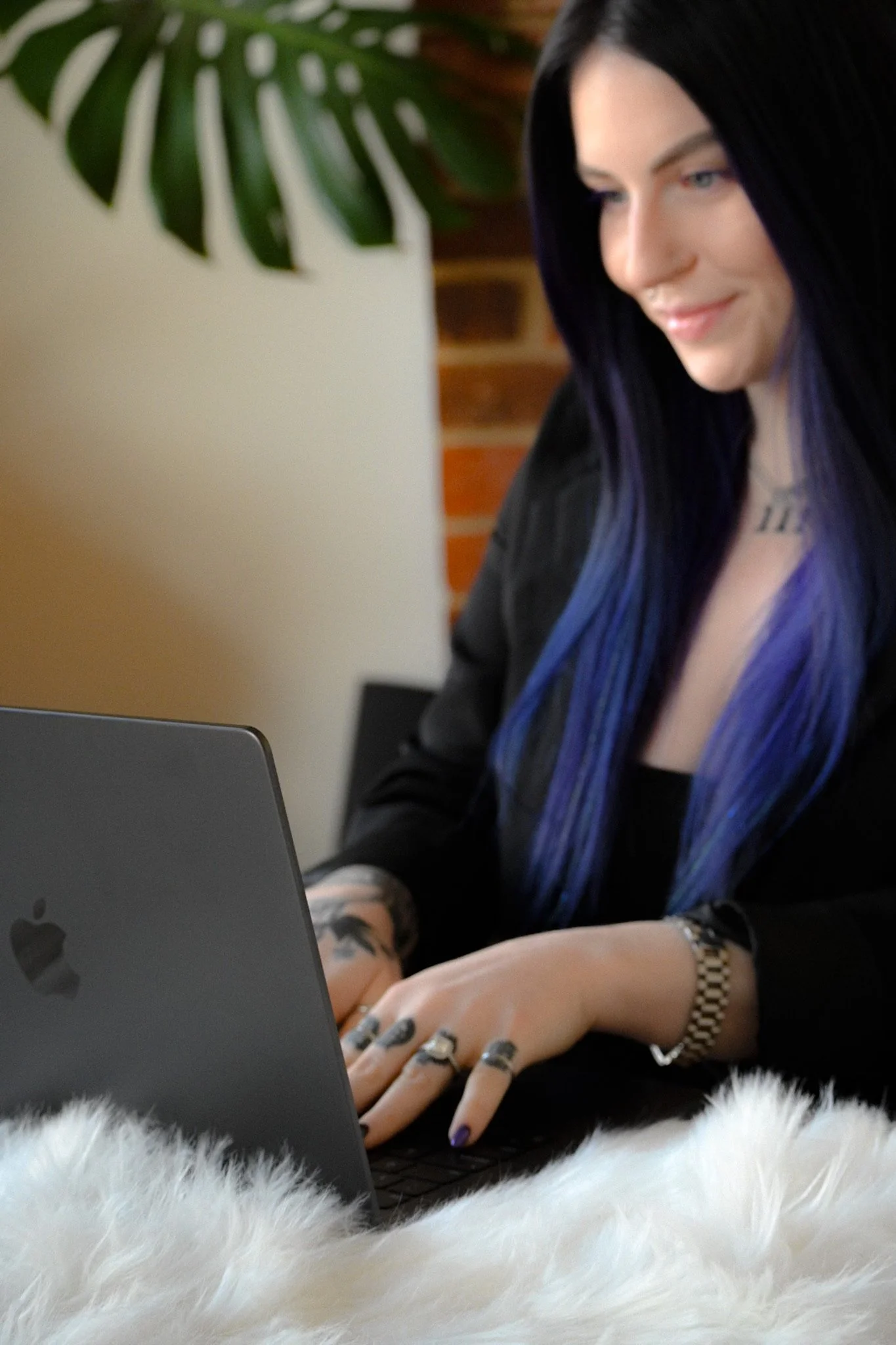 A woman with long black and blue hair working on a laptop at a desk with a white fluffy surface, a plant, and a brick wall in the background.