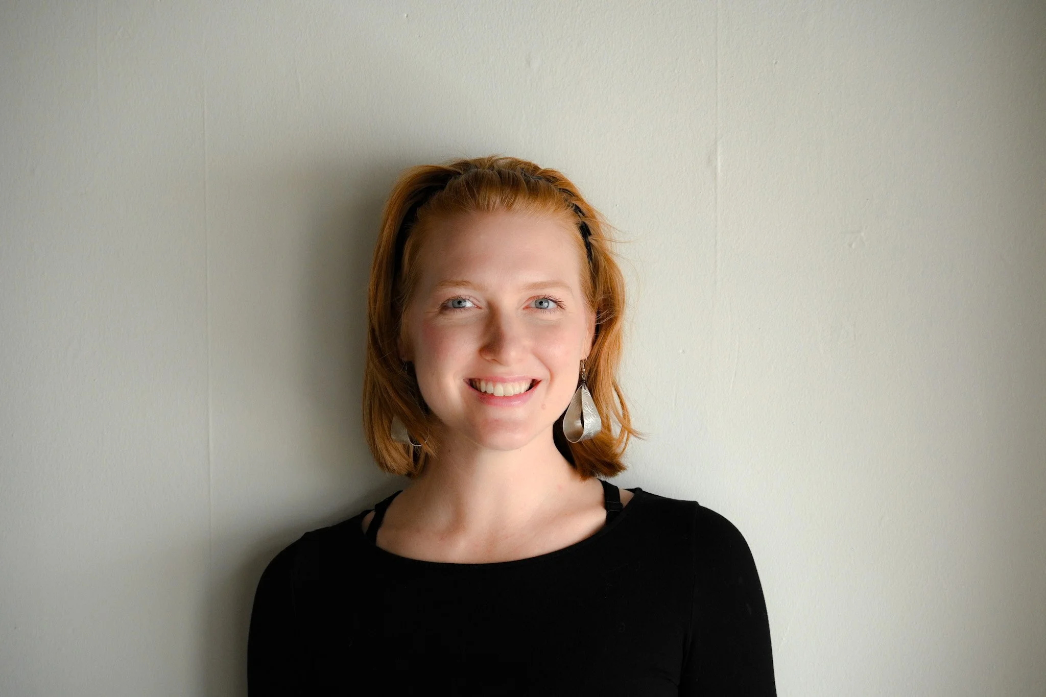 A woman with red hair, wearing large silver earrings, smiling and looking at the camera, standing against a plain light-colored wall.