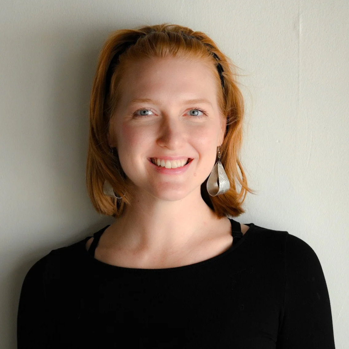 A woman with short red hair and blue eyes, wearing a black top and hoop earrings, standing against a plain white wall.