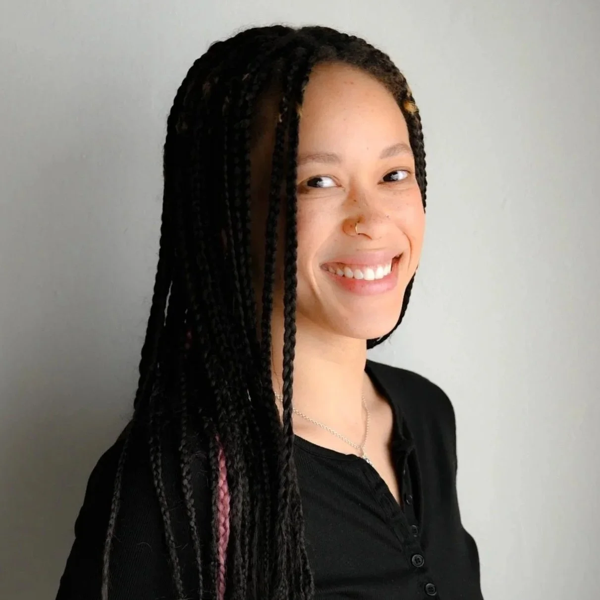 A young woman with long, braided hair smiling at the camera, wearing a black turtleneck sweater and silver jewelry, standing against a plain light-colored background.