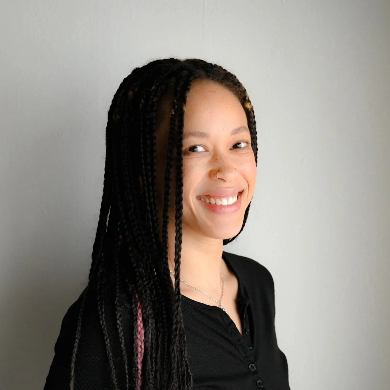 A young woman with long braids, some with pink streaks, smiling and looking to the side, wearing a black top, standing against a plain light gray wall.