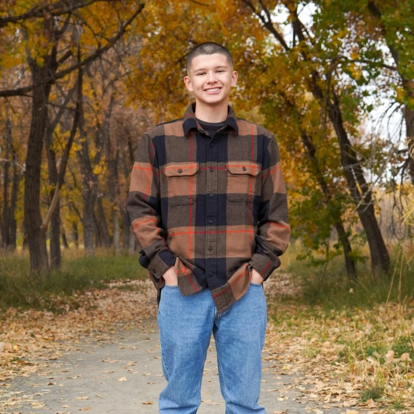 Young man standing on a forest path during autumn, smiling, wearing a plaid jacket and blue jeans.