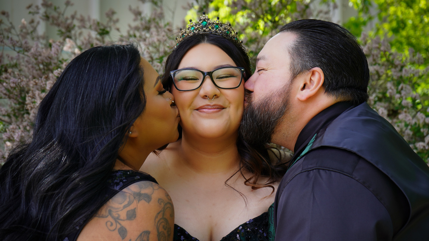 A woman with glasses and a crown is being kissed on each cheek by a man and a woman outside with blooming trees in the background.