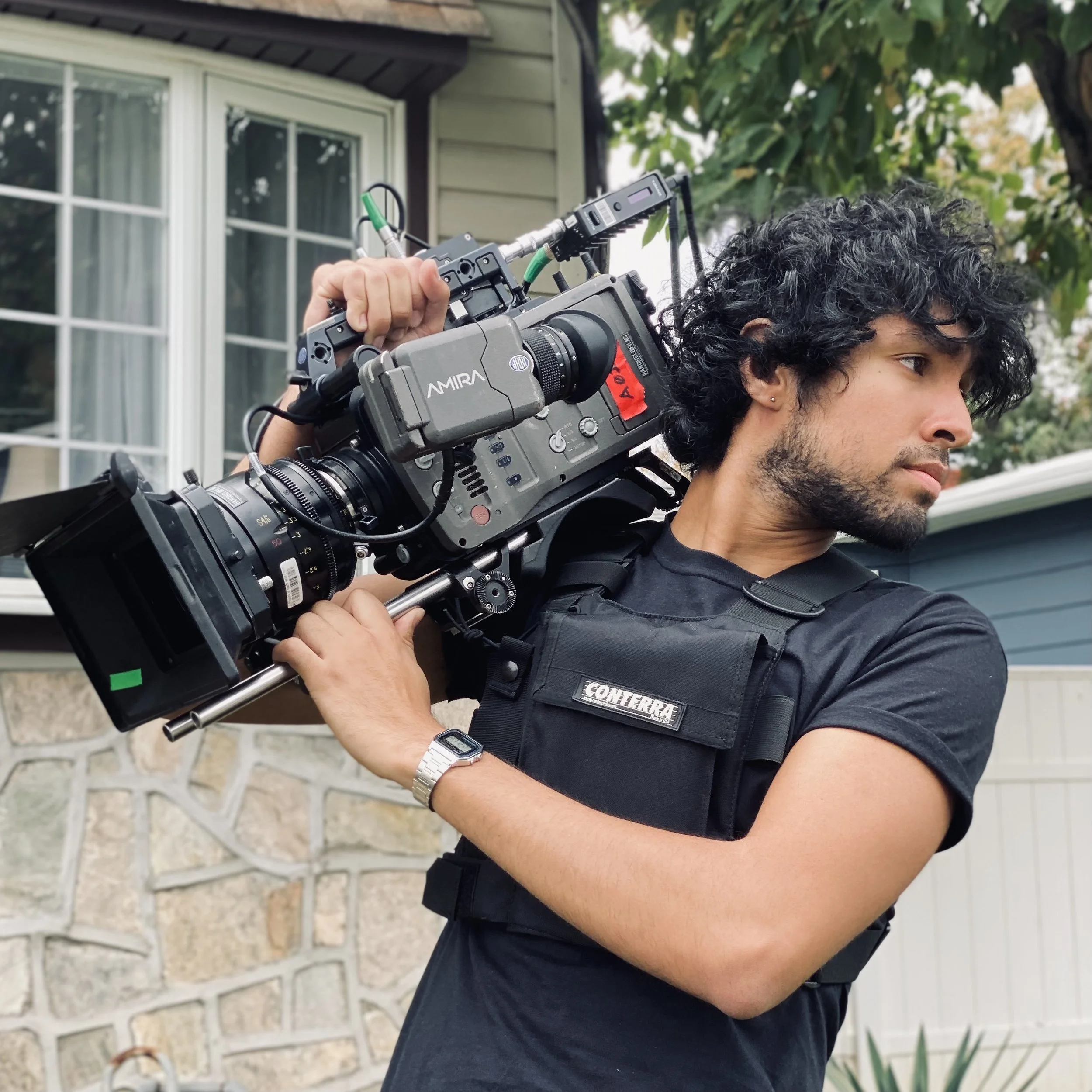 Joao Cueto, A man with curly black hair, wearing a black T-shirt and a black tactical vest labeled 'CONTERRE', is holding a professional video camera on his shoulder outdoors in front of a house with trees and a stone wall.